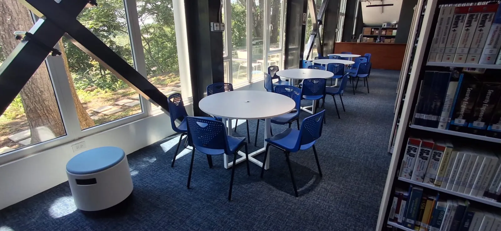 Zona de estudio de una biblioteca universitaria con mesas redondas y sillas azules junto al ventanal